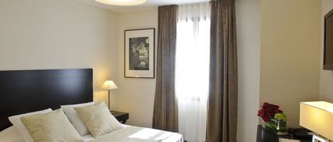 Une chambre d’hôtel avec un grand lit, une table de chevet, une lampe, un tableau encadré au mur et une fenêtre avec des rideaux.
