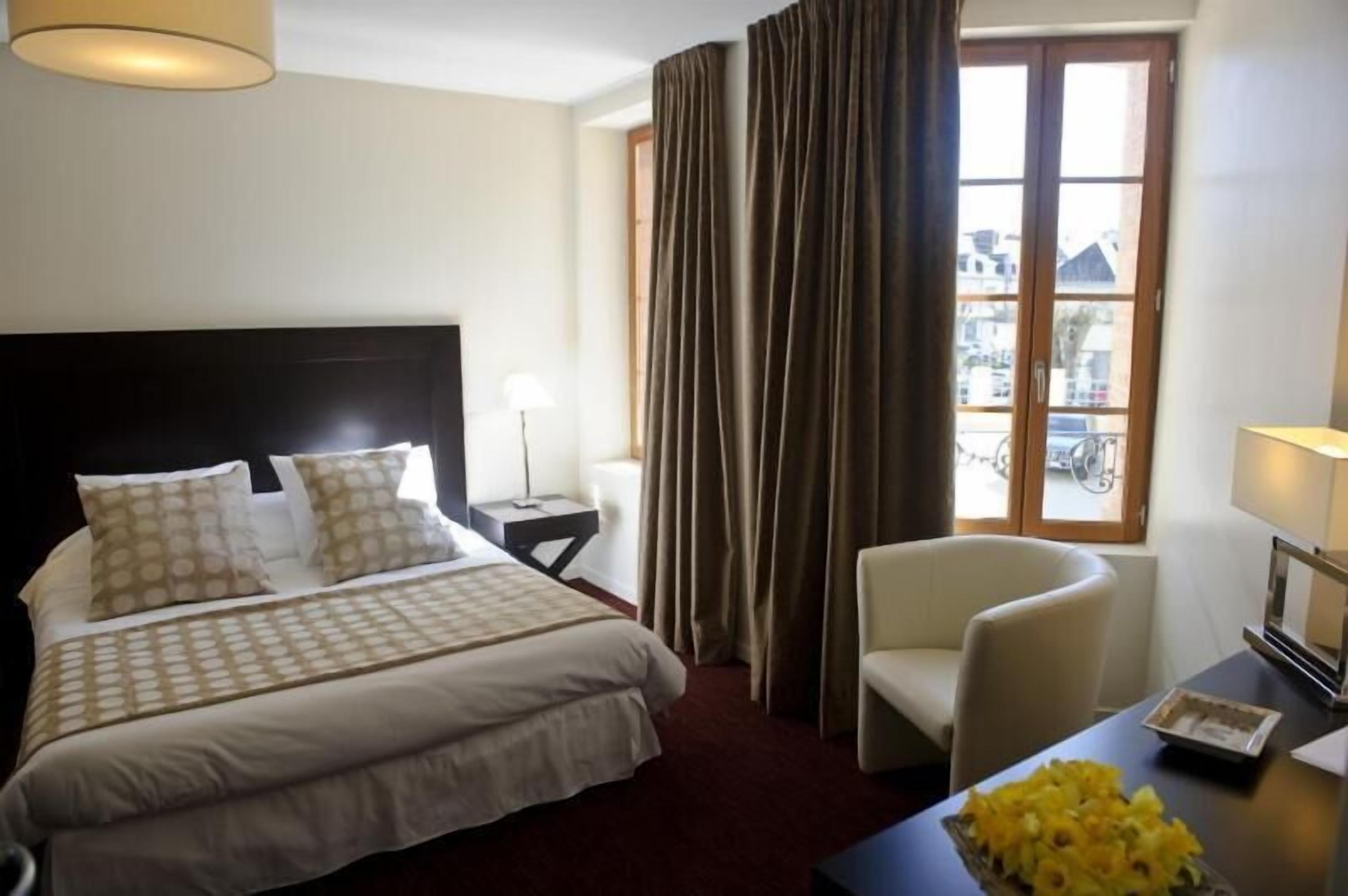 Una camera d'albergo con un letto grande, una sedia, un tavolino con una lampada e una finestra con tende.