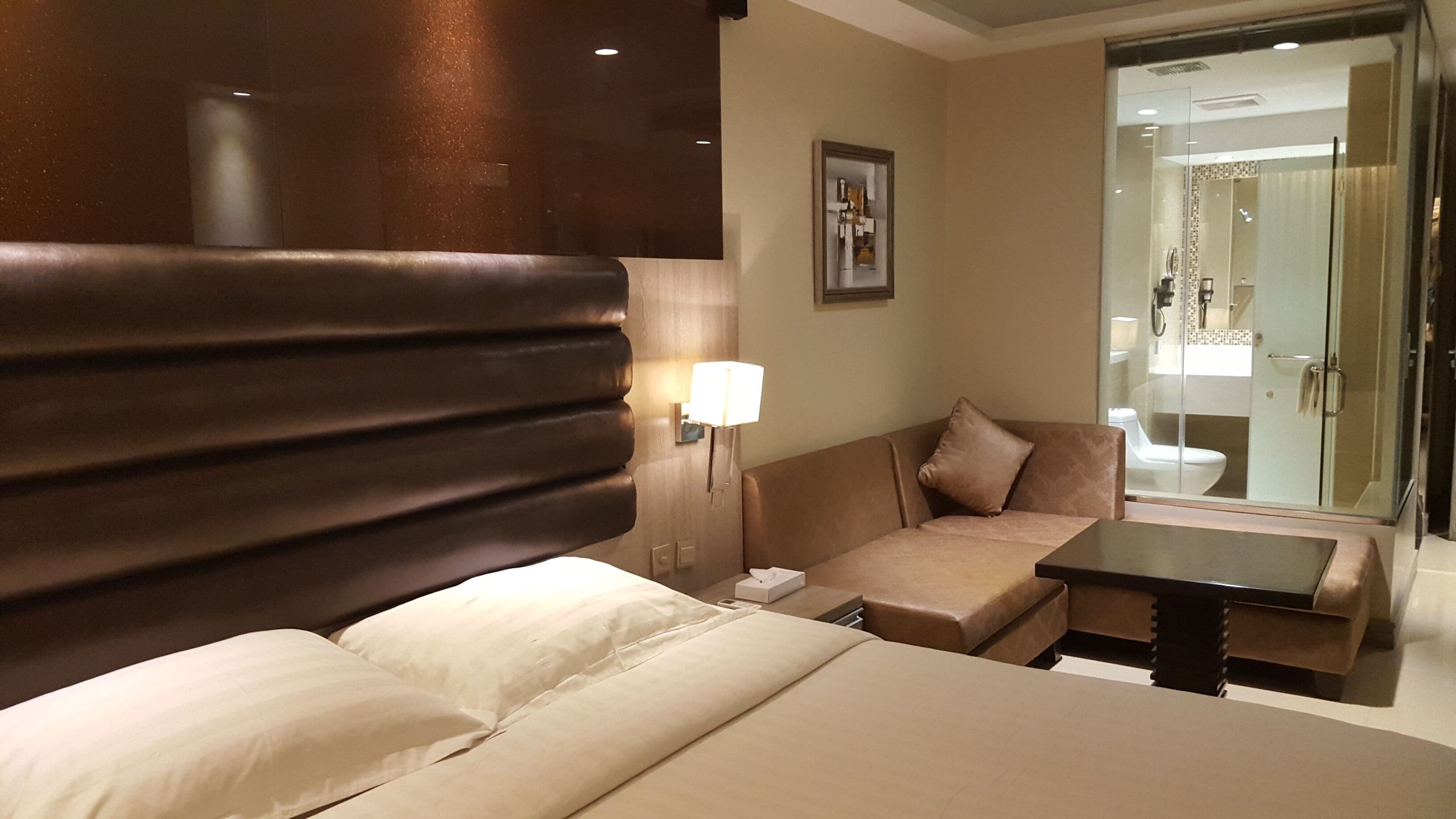A hotel room with a large bed, a sofa, a small table, and a bathroom visible through a glass door.