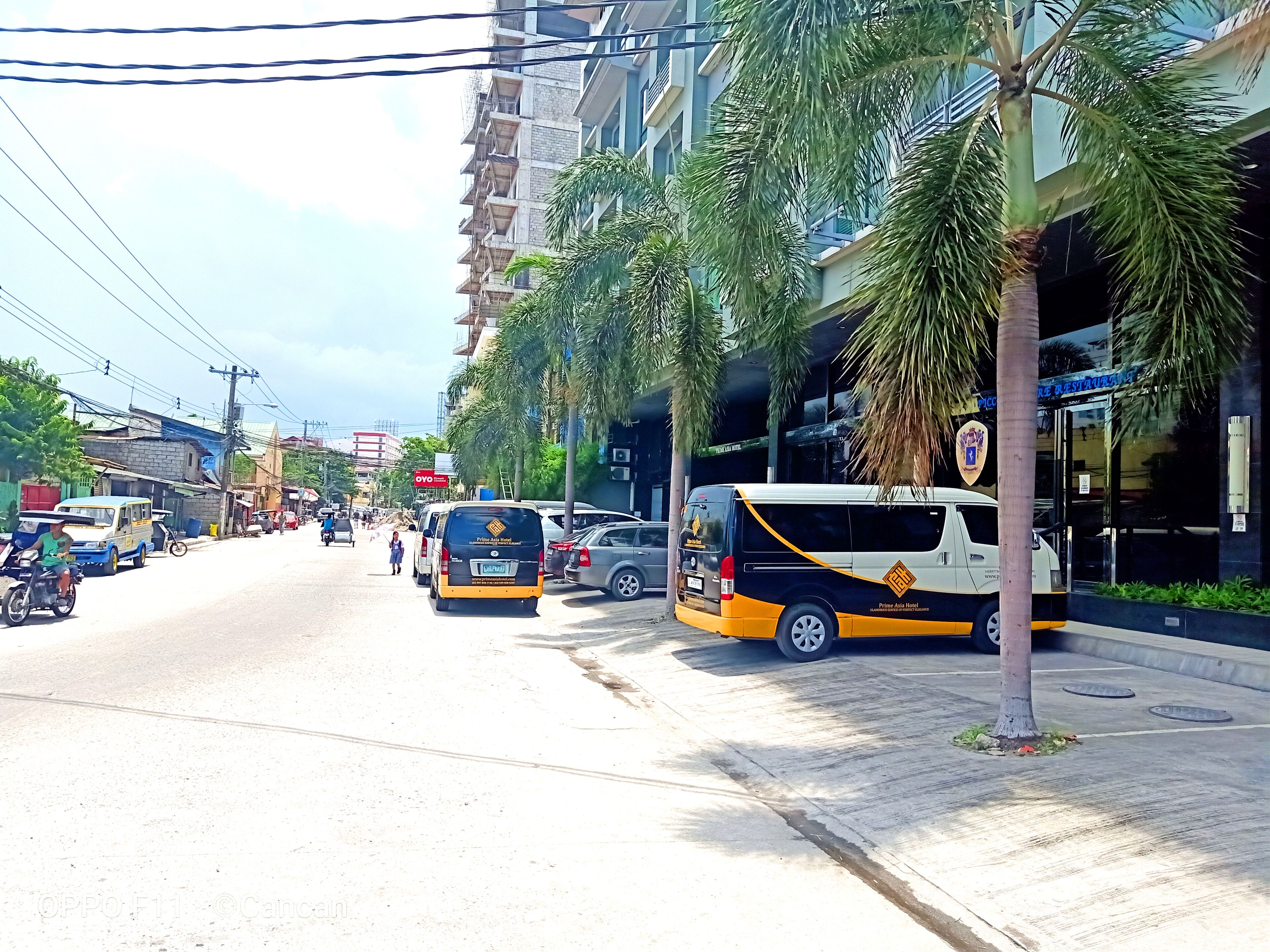 A street with a building that has a sign for 'OYO' and a 'Prime Asia Hotel' sign. There are vehicles parked along the street, including a van with a yellow and black design.