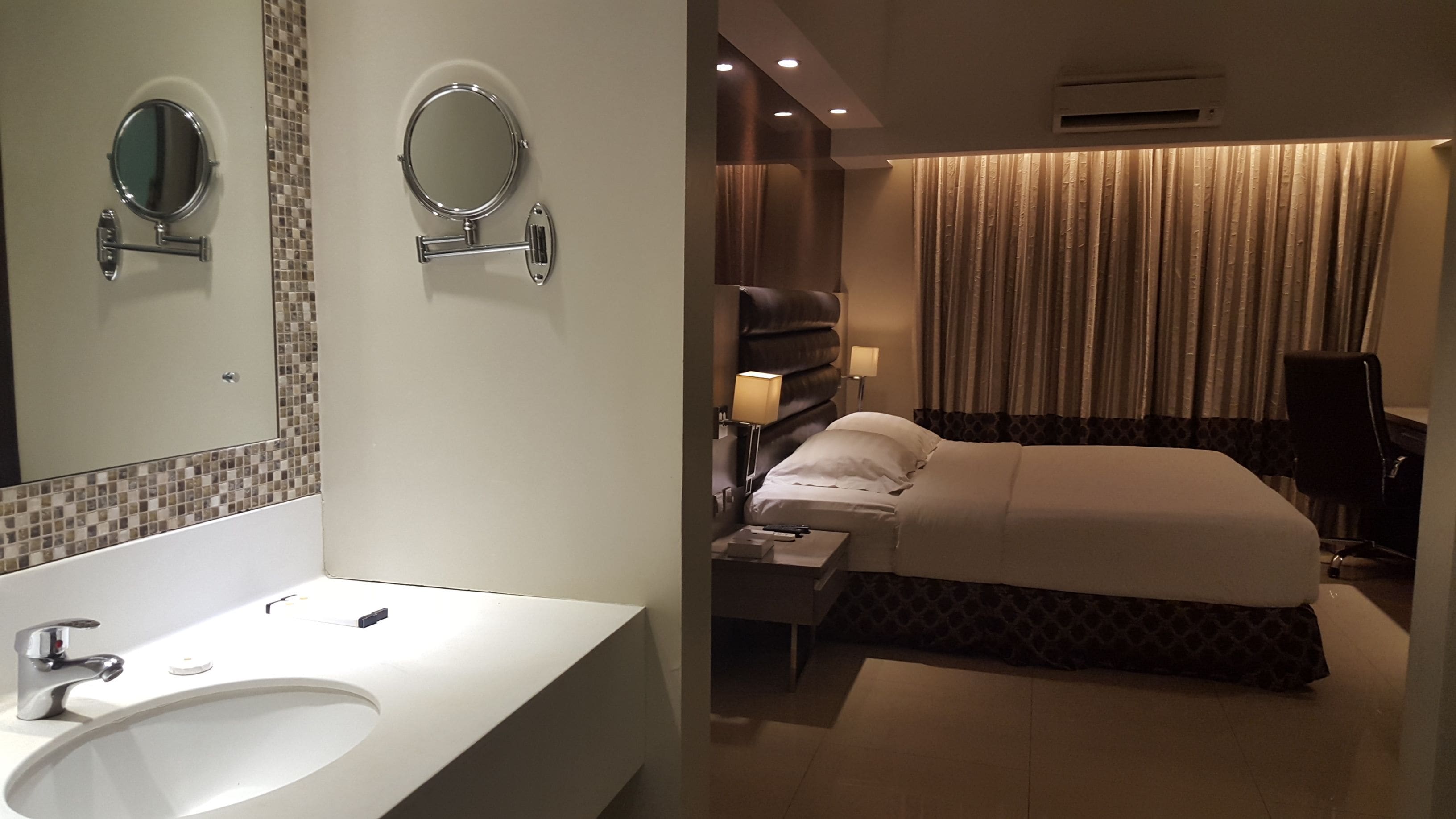 Une chambre d’hôtel avec un grand lit, une table de chevet avec une lampe, un miroir et une salle de bain équipée d’un lavabo et de deux miroirs ronds.