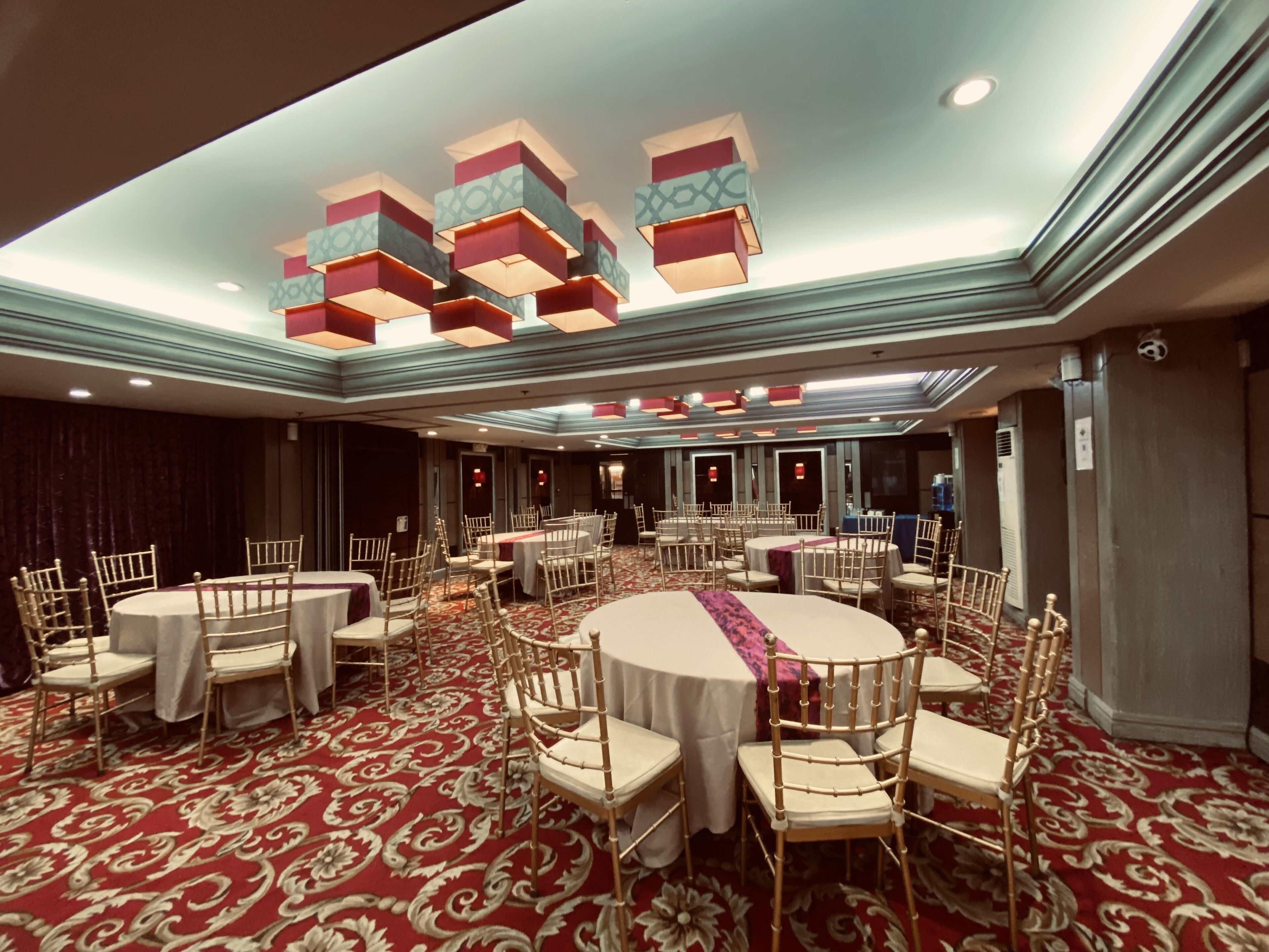 A banquet hall with round tables and chairs, a patterned carpet, and a ceiling with geometric light fixtures.