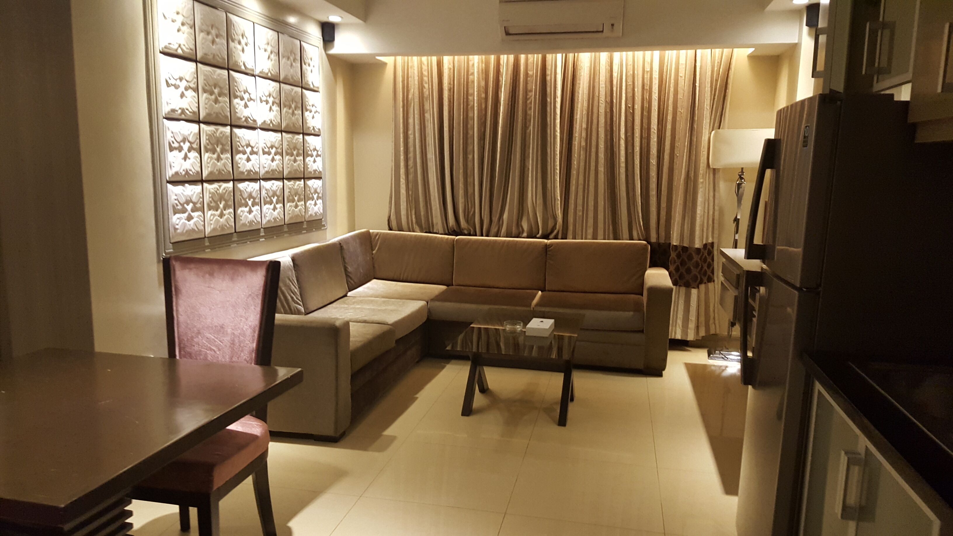 A modern living room with a sectional sofa, a glass coffee table, a dining table with chairs, a refrigerator, and a decorative wall panel.
