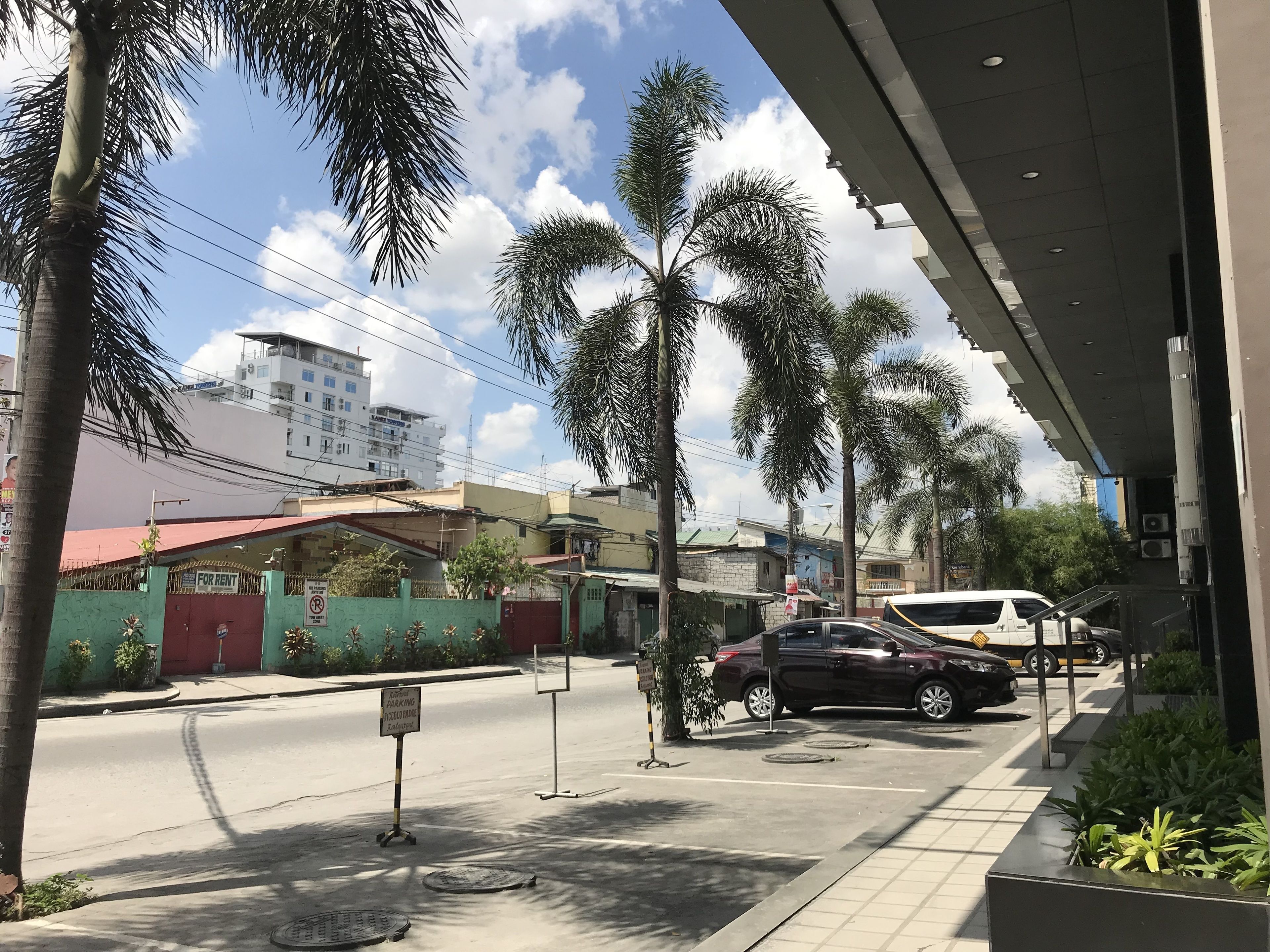 A street view with parked cars, a building with a red roof, and palm trees.