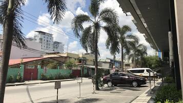 A street view with parked cars, a building with a red roof, and palm trees.