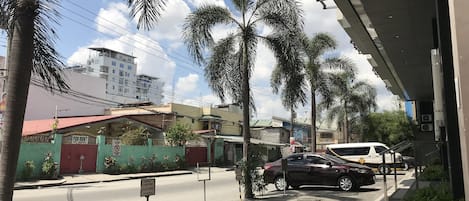 A street view with parked cars, a building with a red roof, and palm trees.