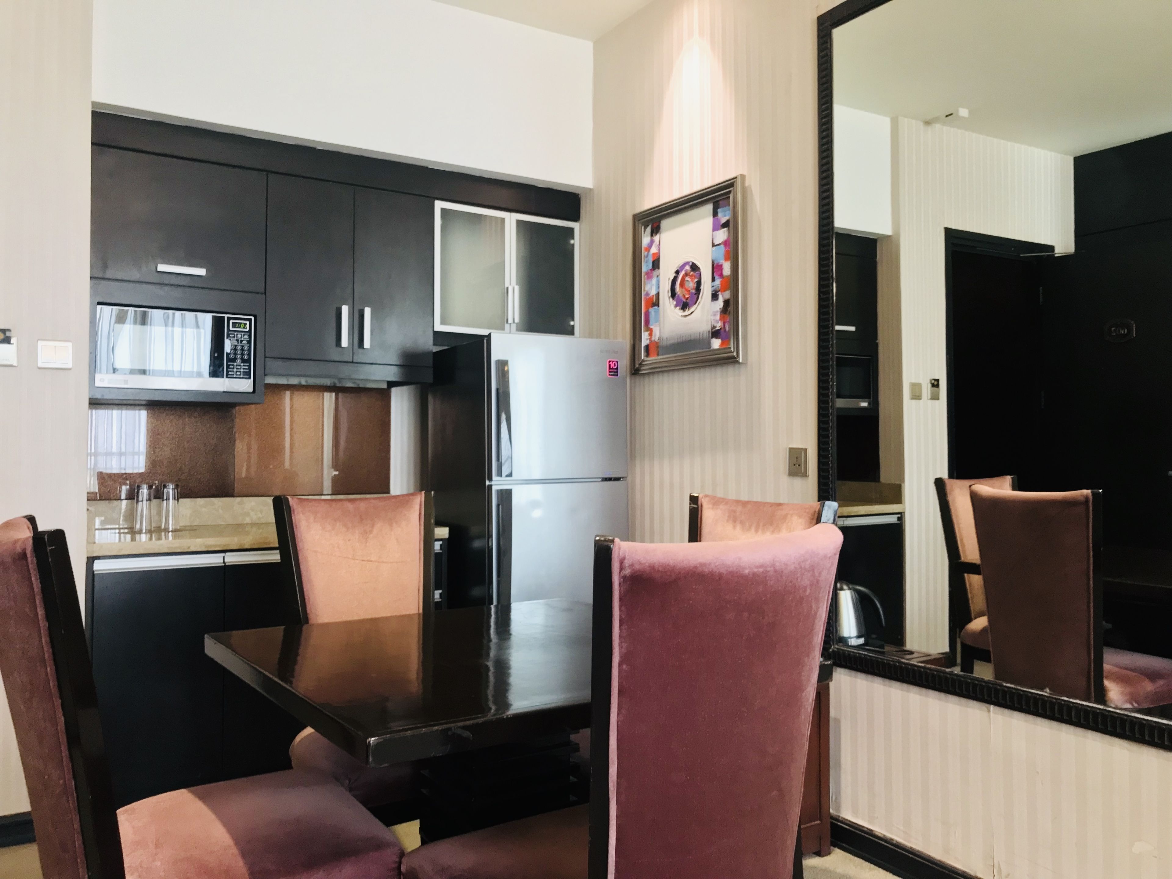 A compact kitchen with dark cabinets, a stainless steel refrigerator, and a microwave. There is a dining table with chairs, and a large mirror on the wall.