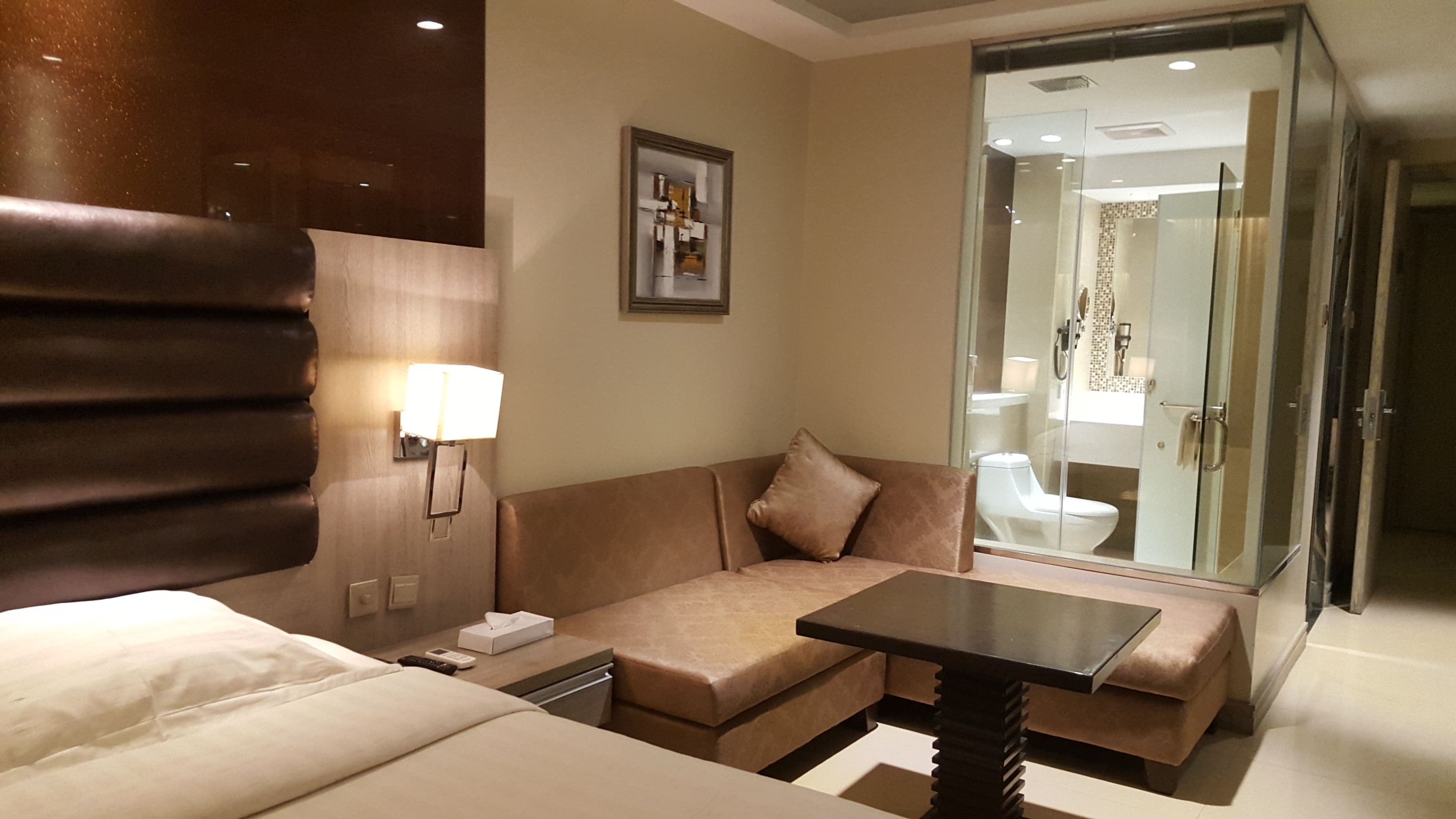 A hotel room with a bed, a sofa, a table, a lamp, a picture on the wall, and a glass door leading to a bathroom.