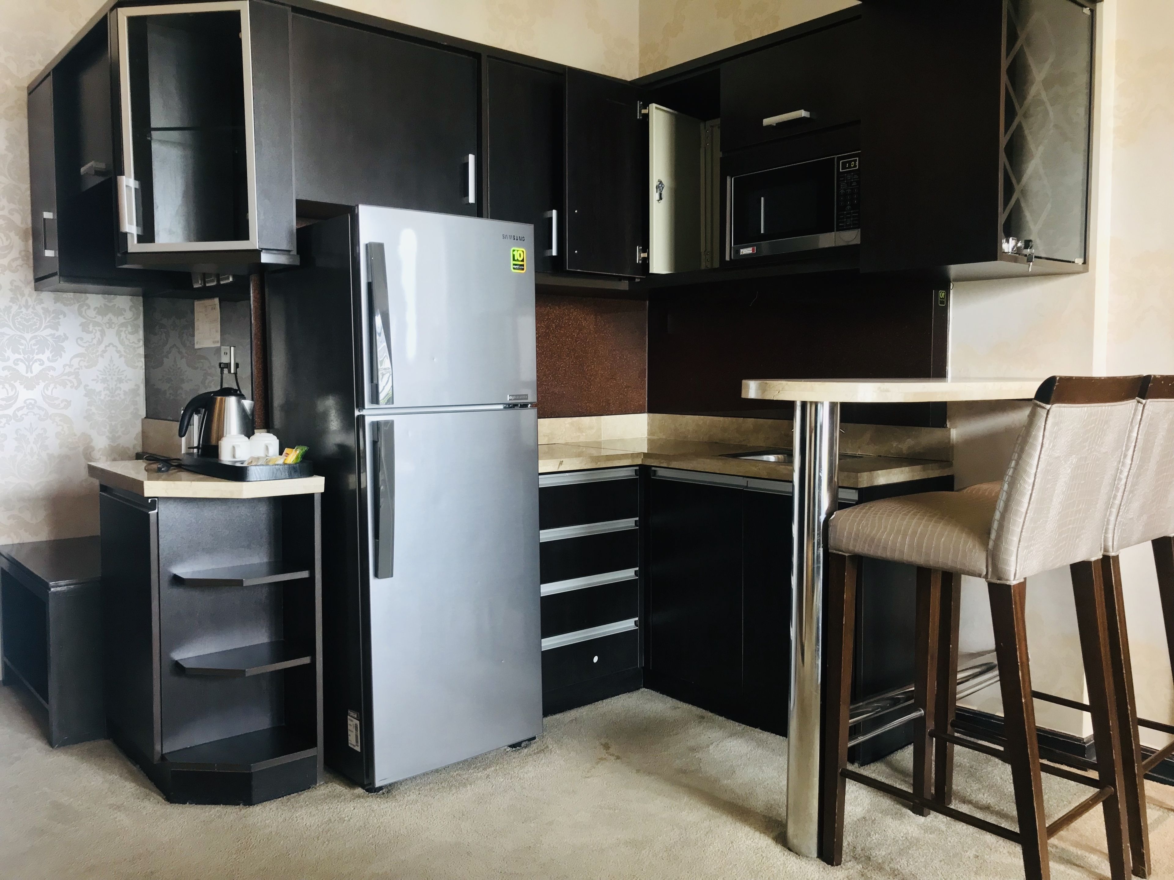 Una cocina compacta con un refrigerador de acero inoxidable, un microondas y un taburete pequeño.
