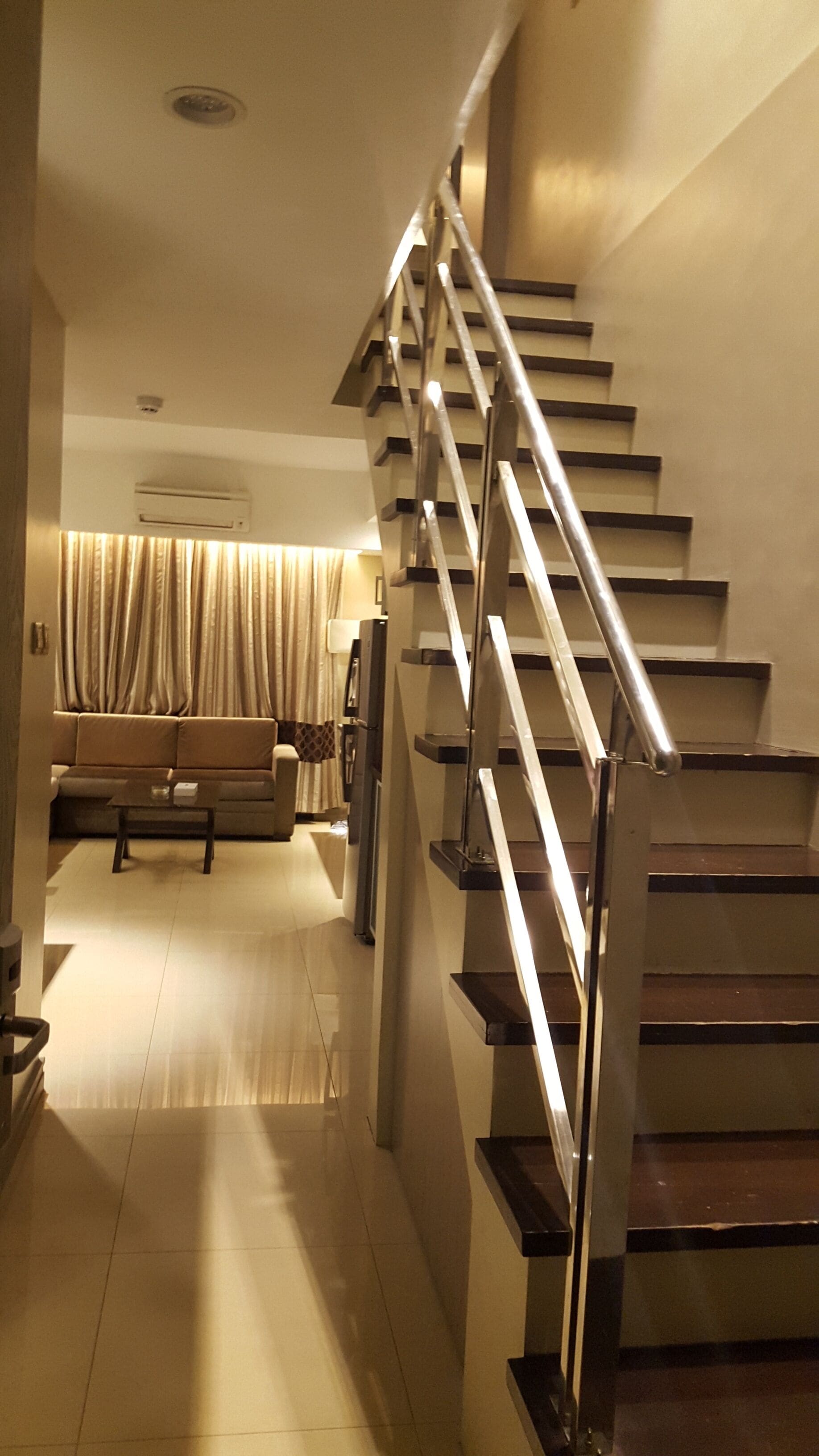 Un escalier moderne avec une rampe en métal, menant à un espace de vie comprenant un canapé et des rideaux.