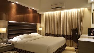 A hotel room with a large bed, a desk with a chair, a lamp, and a window with curtains.