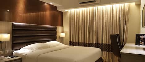 A hotel room with a large bed, a desk with a chair, a lamp, and a window with curtains.
