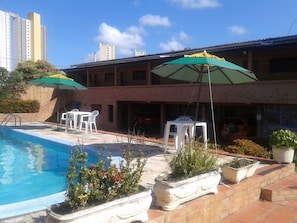 Una piscina con sedie bianche e ombrelloni verdi, circondata da piante in vaso e un edificio in mattoni.