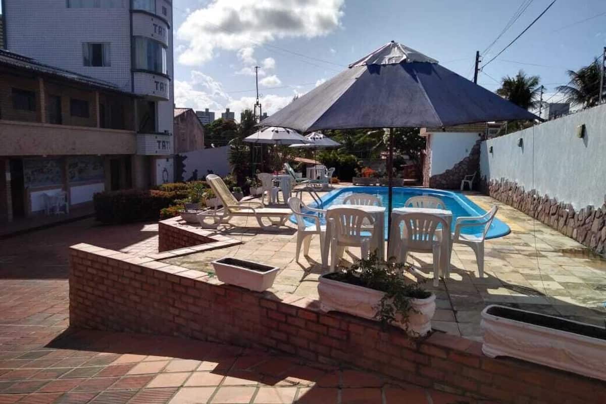 A pool area with lounge chairs and umbrellas, surrounded by a brick wall and a building with balconies.