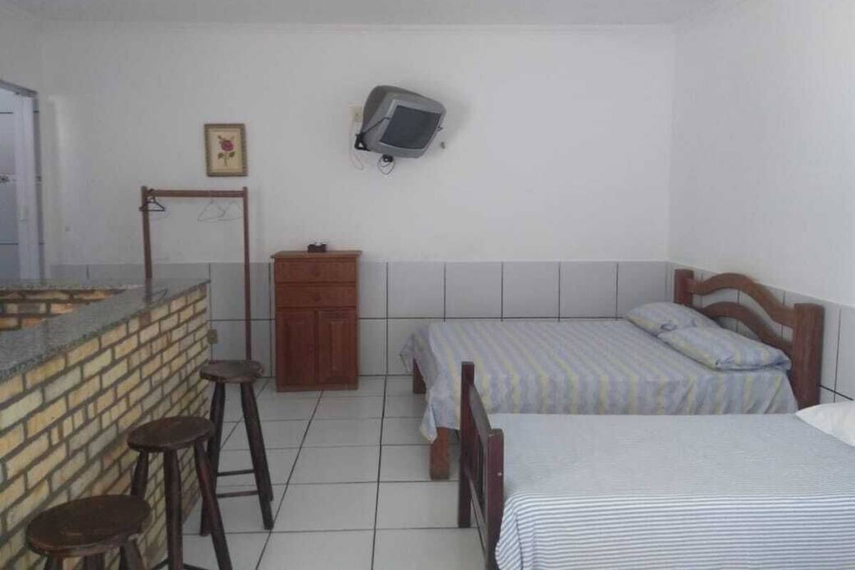 A small, clean room with a kitchenette, two single beds, a wooden cabinet, and a television mounted on the wall.
