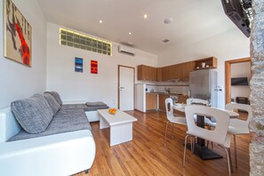 A modern living room with a white sofa, a coffee table, and a dining area with white chairs. A kitchen with wooden cabinets and a refrigerator is visible in the background. - Marmontova Luxury Rooms (Split)
