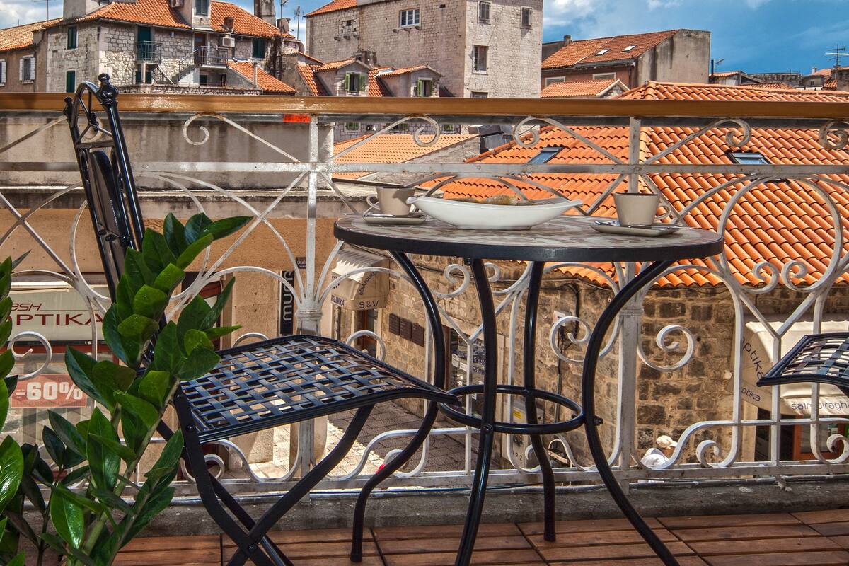 A terrace with a table and chairs, a potted plant, and a view of buildings with red-tiled roofs.