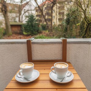 Två vita kaffekoppar står på ett träbord tillsammans med små fat och teskedar. Bordet är placerat på en balkong med utsikt över träd och byggnader.