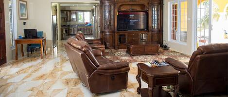 A spacious living room with a large leather sofa, a wooden entertainment center, and a dining area visible through an open doorway.