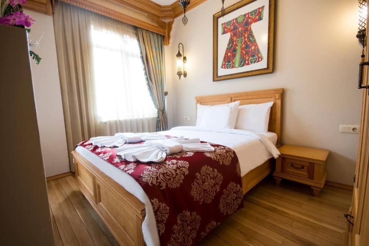 Habitació d'hotel amb un llit de fusta, un llençol de llit vermell i blanc, una petita taula de nit de fusta i una làmpada penjada a la paret.