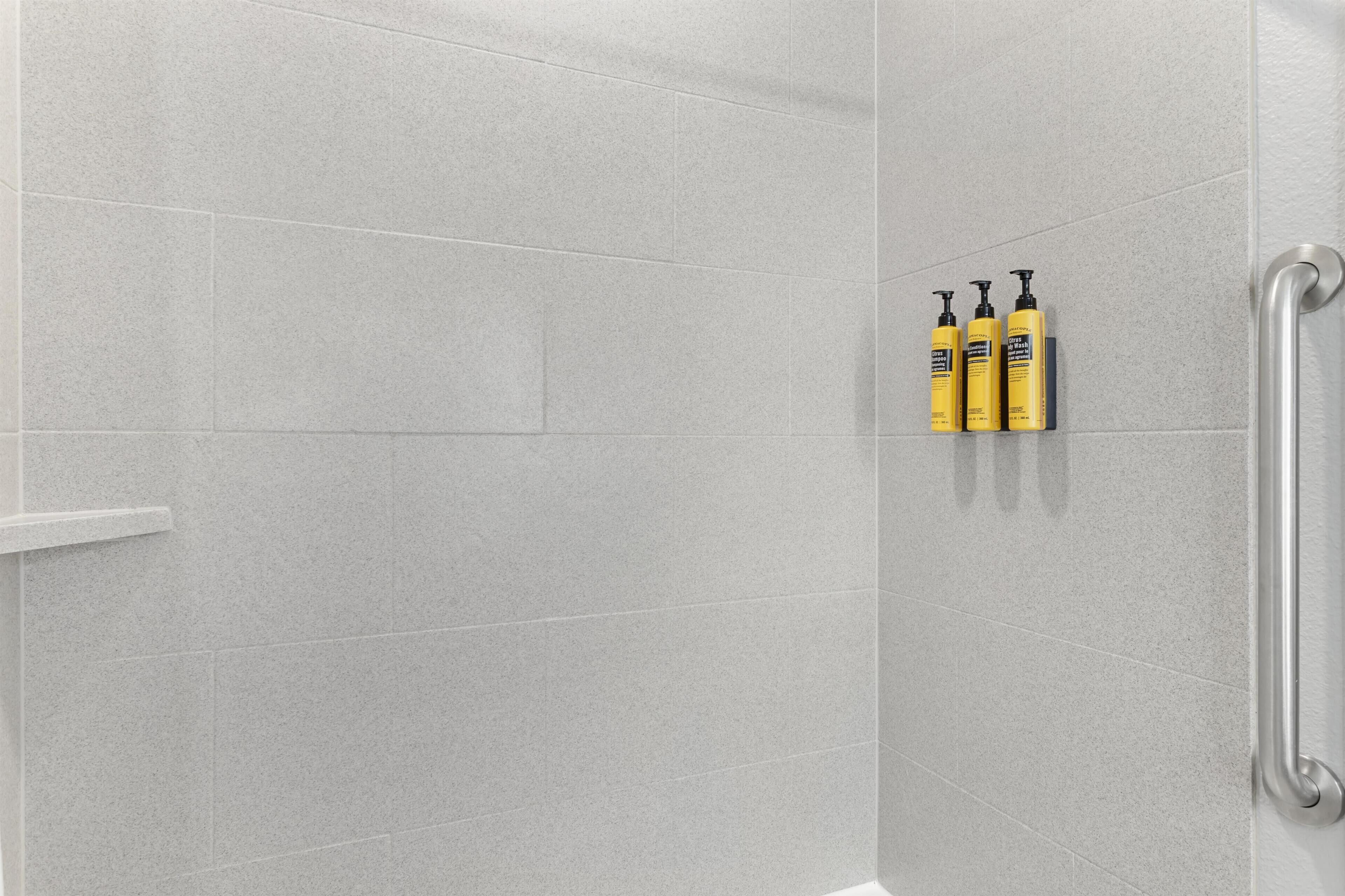 Un angolo doccia con piastrelle grigie, una barra di sostegno bianca e tre flaconi di shampoo gialli.