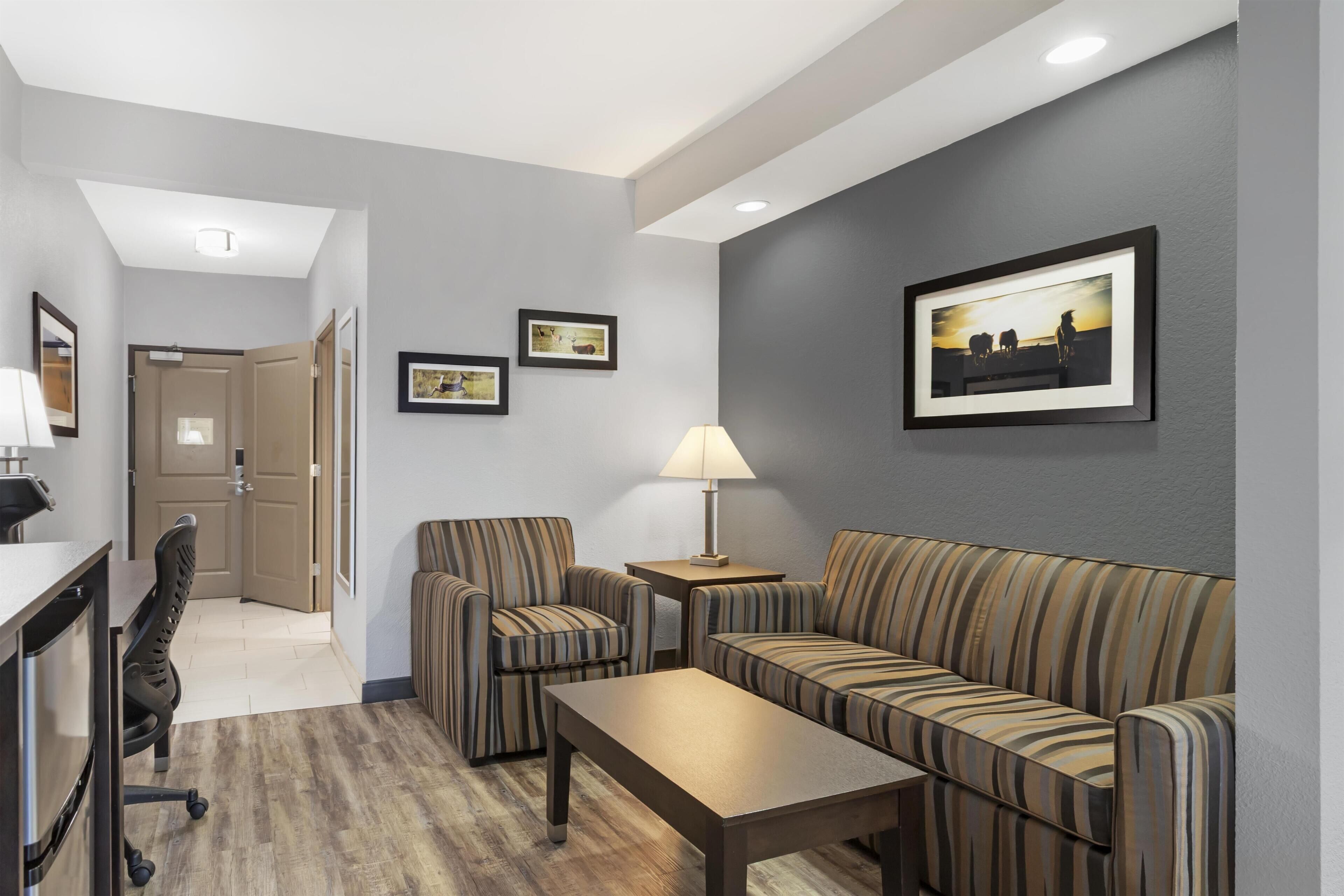 A hotel room with a sofa, armchair, and desk. There are framed pictures on the wall and a lamp on the side table.
