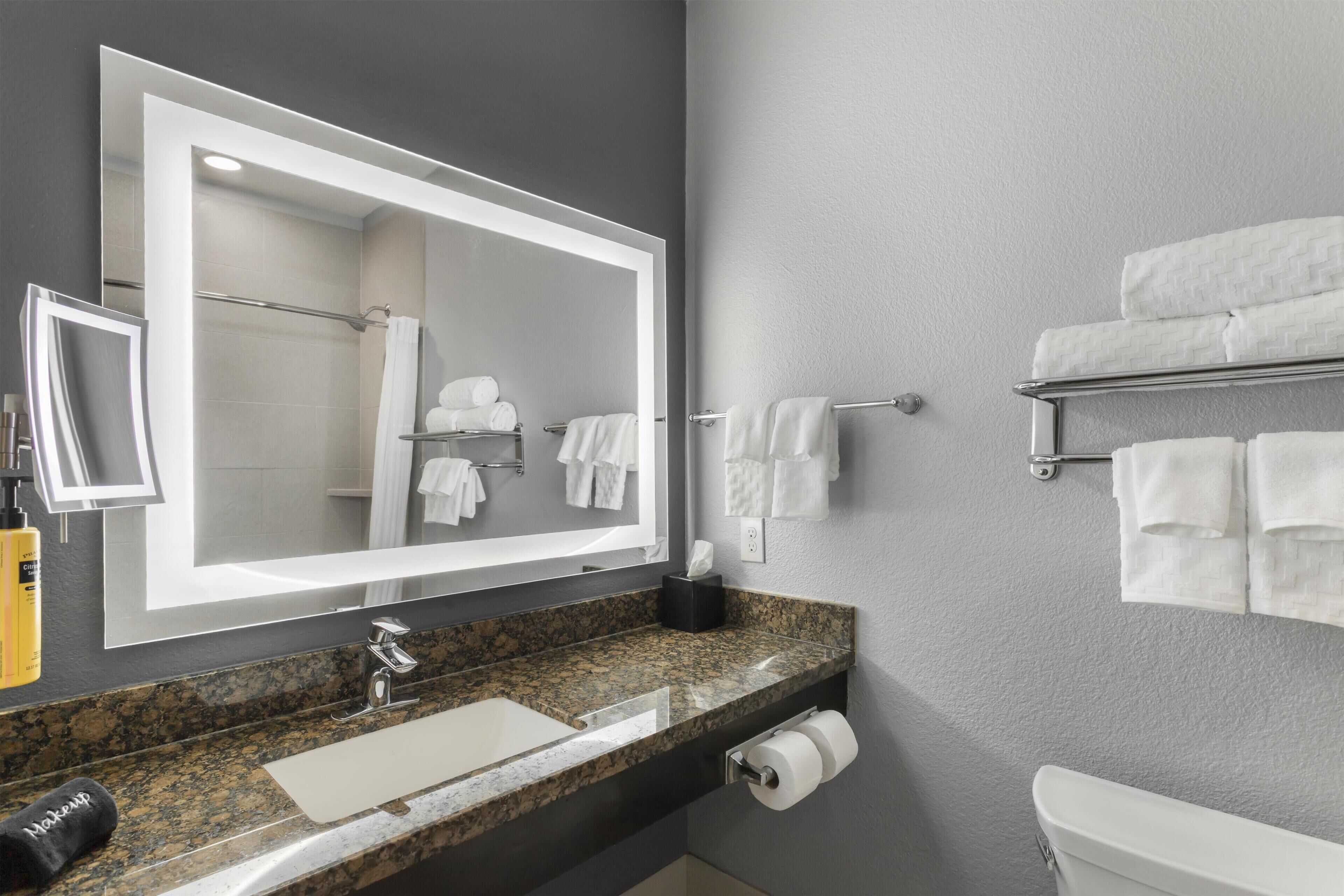 Un baño moderno con un espejo grande, encimera de granito, lavamanos, inodoro y toalleros.