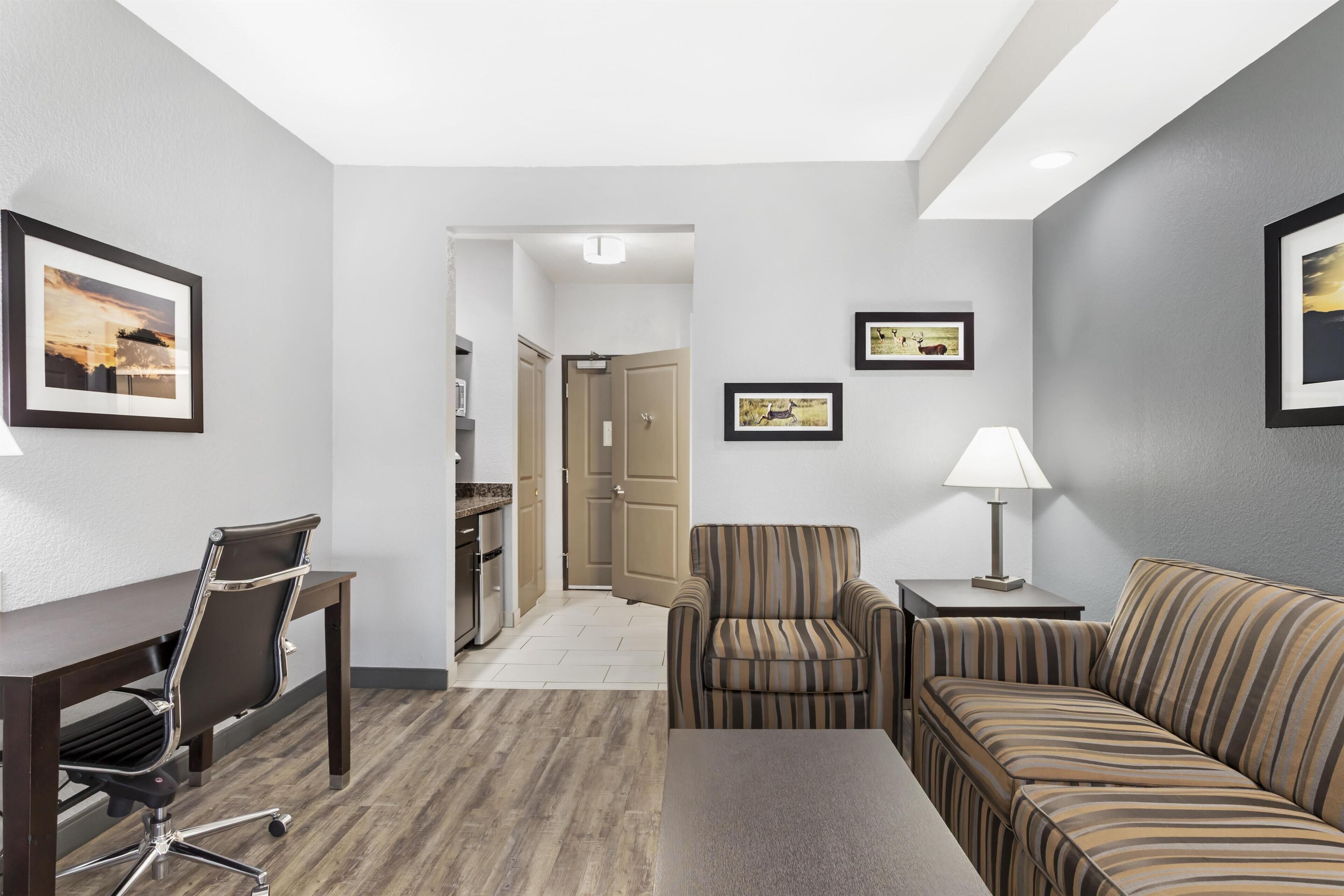 A hotel room with a desk, chair, sofa, and a small table with a lamp. There are framed pictures on the walls.
