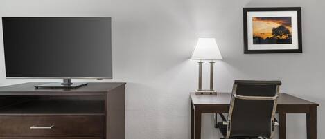 Une chambre d’hôtel équipée d’une télévision à écran plat, d’un bureau avec une lampe, d’un tableau encadré au mur et d’un panier pour animaux de compagnie.