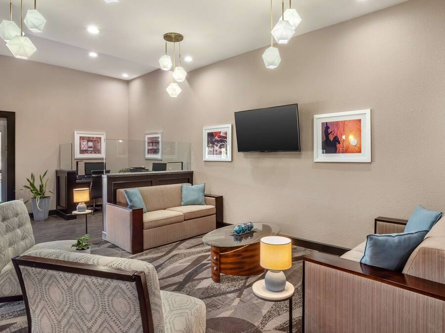 A hotel room with a sofa, armchair, and a small table with a lamp. There is a TV mounted on the wall and a window with a view of the outside.