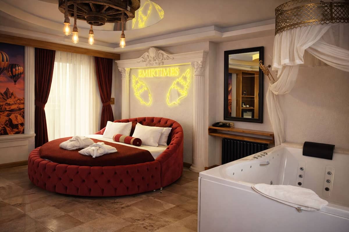 A luxurious bedroom with a red upholstered bed, a large white bathtub, and a neon sign reading "EMIRTIMES."