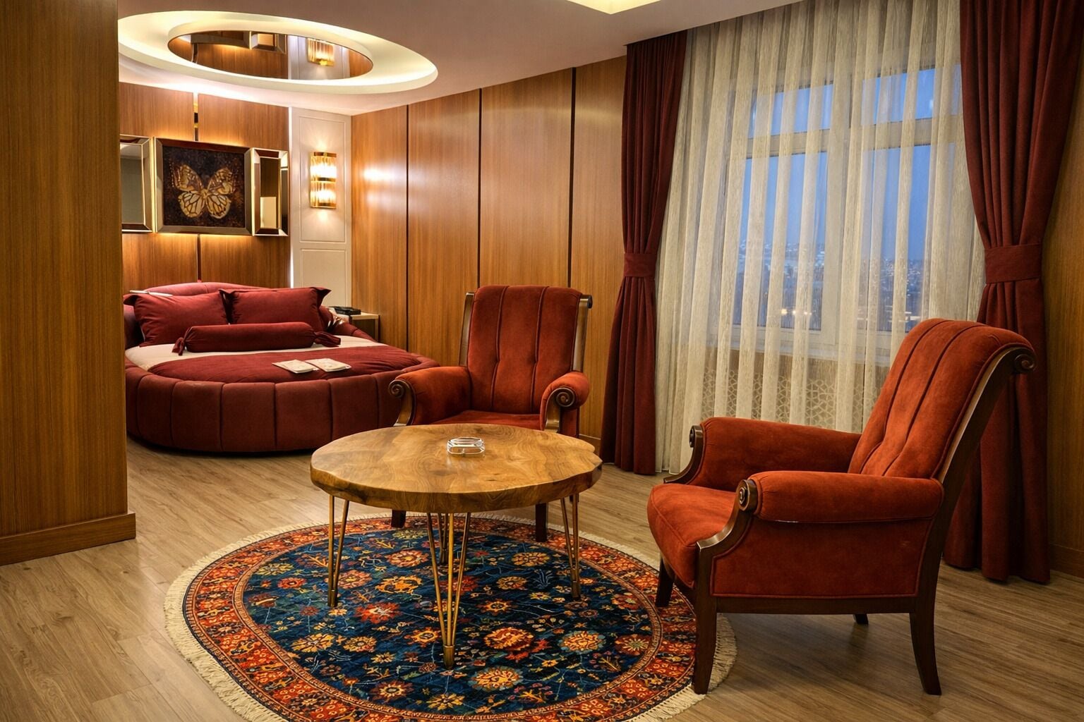 A room with a round wooden table, two red armchairs, a red sofa, a patterned rug, and a large window with sheer curtains.