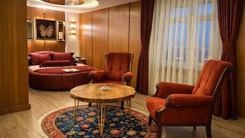 A room with a round wooden table, two red armchairs, a red sofa, a patterned rug, and a large window with sheer curtains.