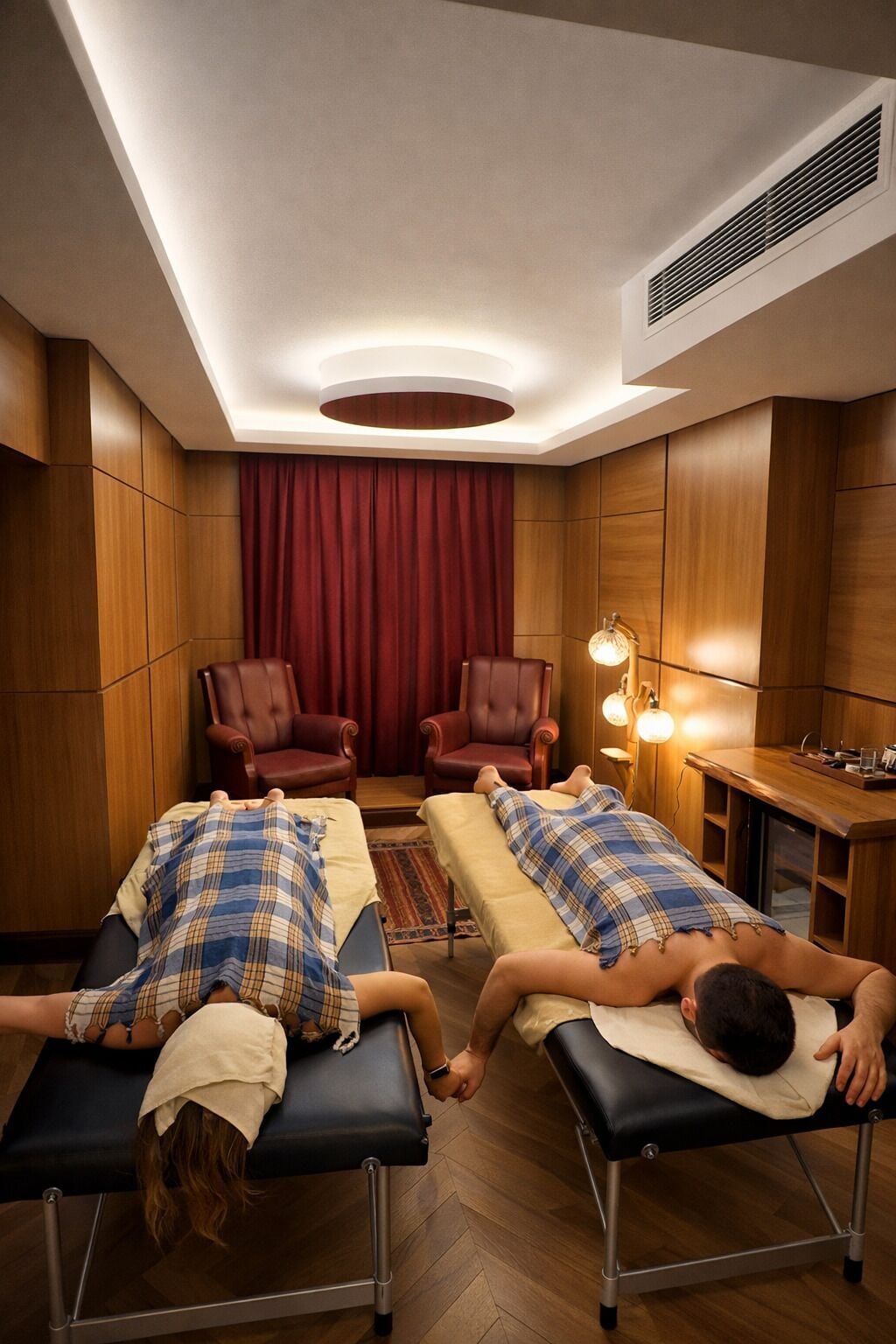 A room with two massage tables, wooden paneling, a red curtain, and a desk with a lamp.