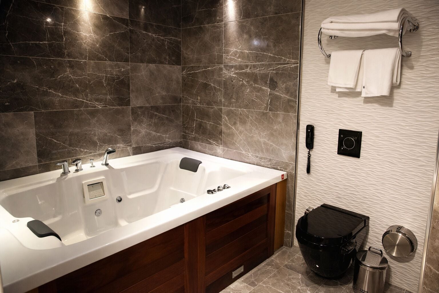 A modern bathroom with a large white bathtub, wooden vanity, marble tiles, a towel rack, a black trash bin, and a wall-mounted phone.