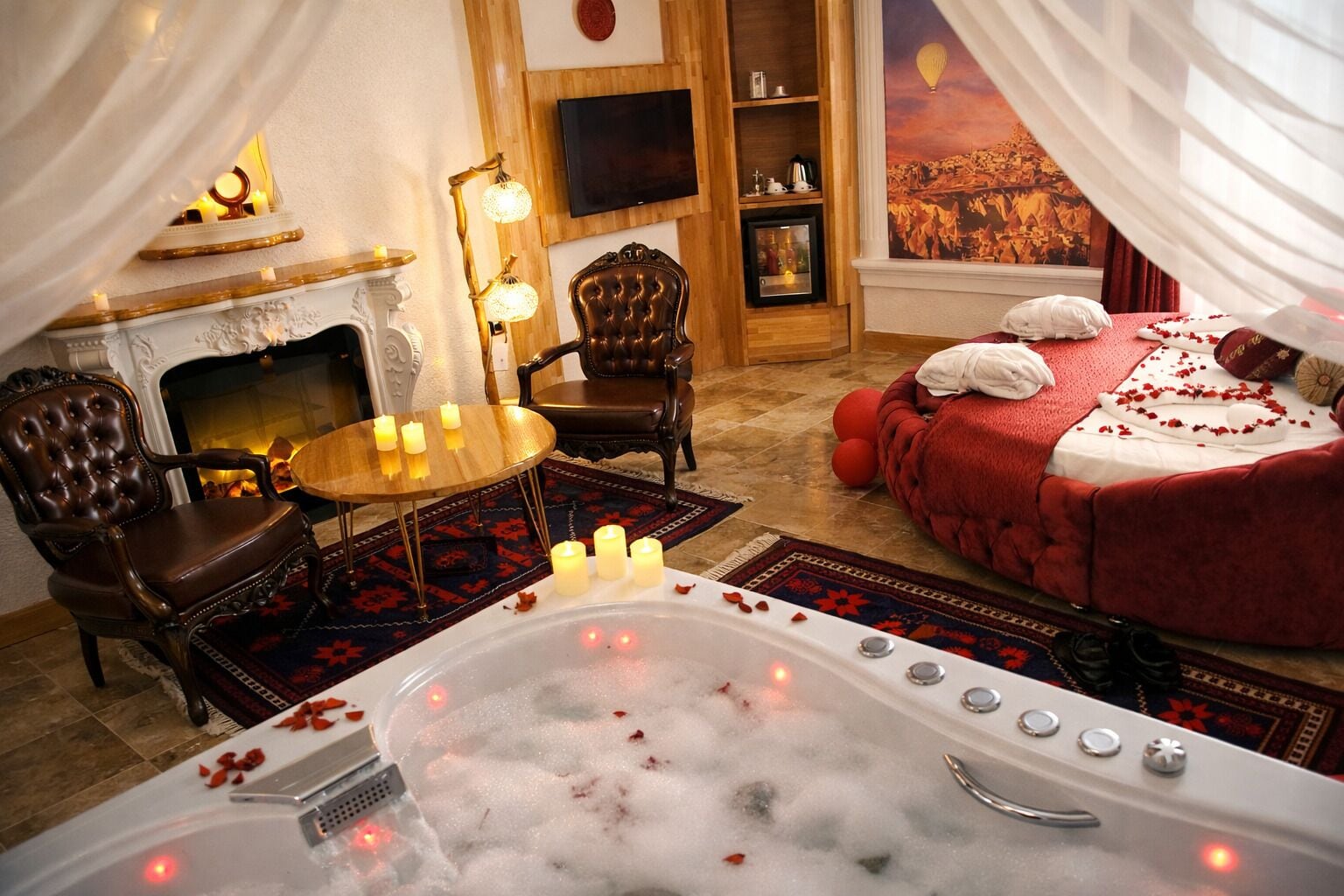 A luxurious bedroom with a large bed, a fireplace, a sitting area with chairs, a bathtub with rose petals, and a television on the wall.