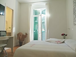 Una habitación de hotel con cama, escritorio, silla, un florero con flores y una ventana con contraventanas.