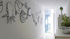 A hallway with a white wall adorned with black line drawings of various sea creatures. There is a door leading to a bright room with a view of the outside. - Altamarea (La Spezia)
