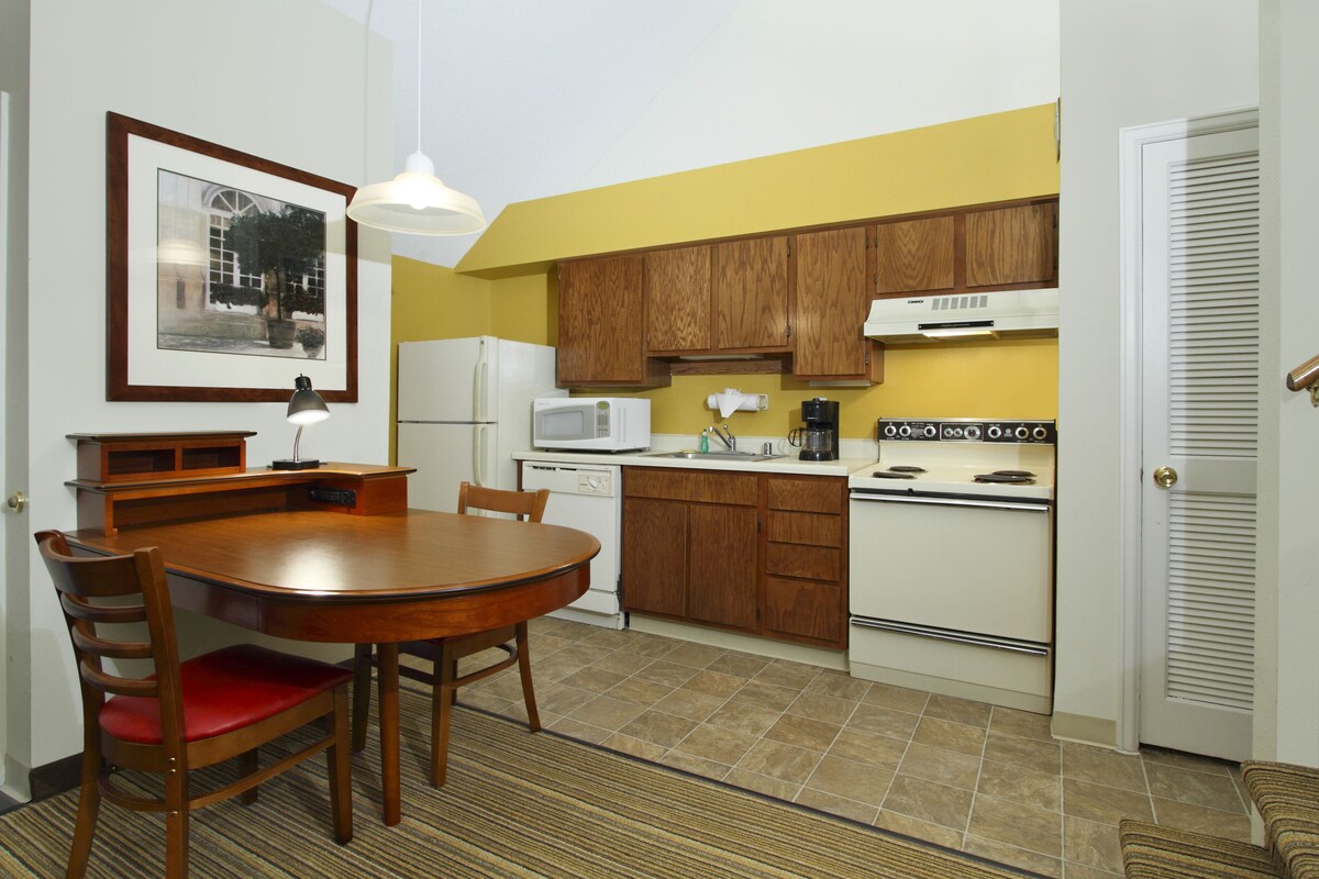 A compact kitchen with wooden cabinets, a white refrigerator, a microwave, and a stove. There is a dining table with chairs and a framed picture on the wall.