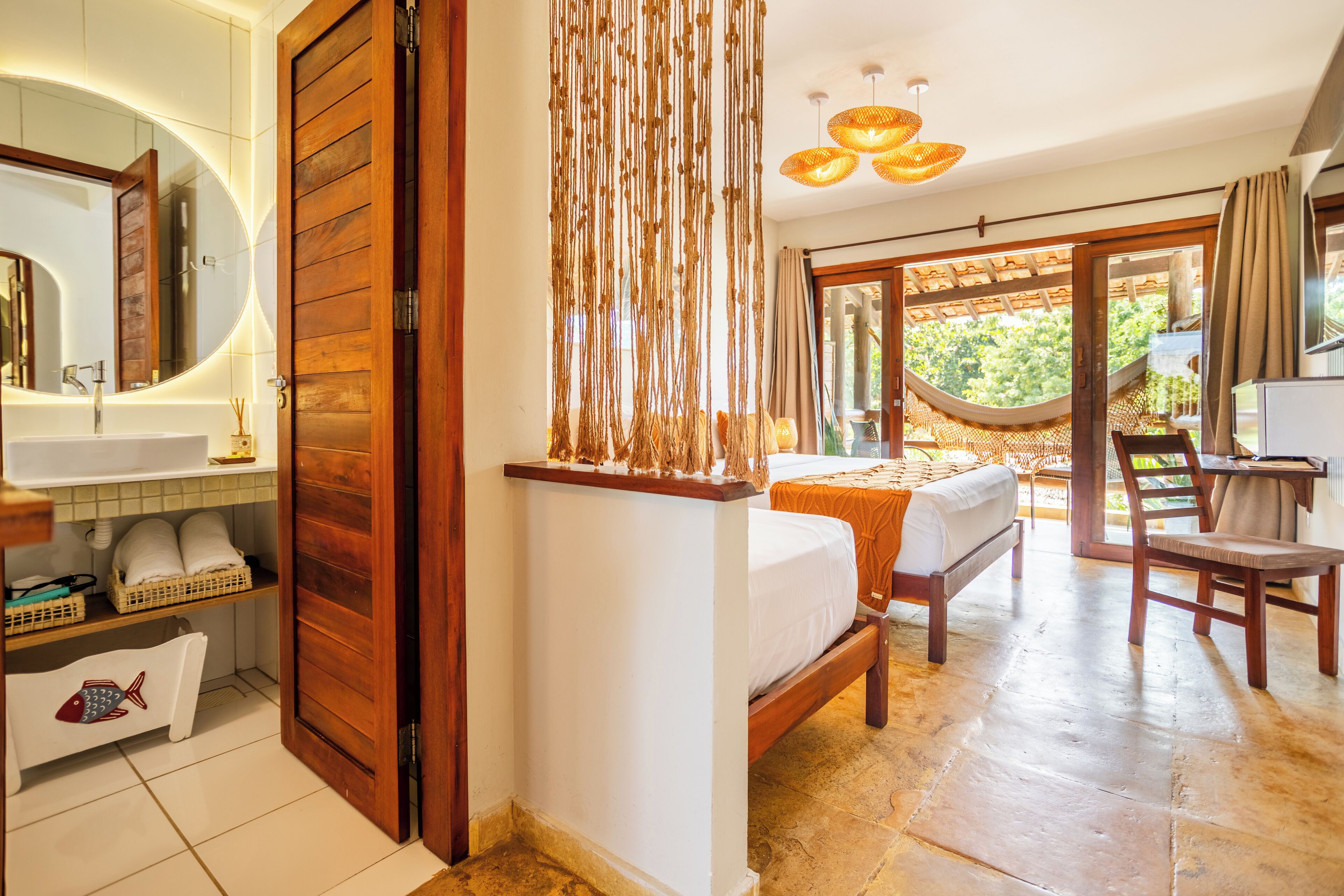 A bedroom with a wooden door, a bed with a white bedspread, a bedside table, a chair, and a view of a hammock and greenery outside.