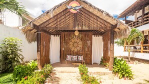 Una estructura con techo de paja y un centro decorativo tejido en el techo, un banco con cojines estampados, y un pequeño jardín con diversas plantas y flores.