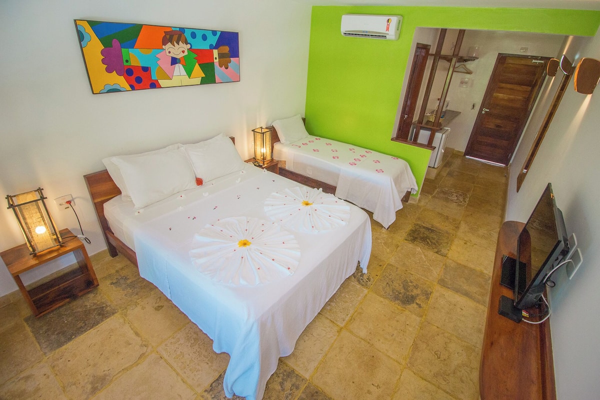 A hotel room with two beds, a small table with a lamp, a painting on the wall, and a green accent wall.