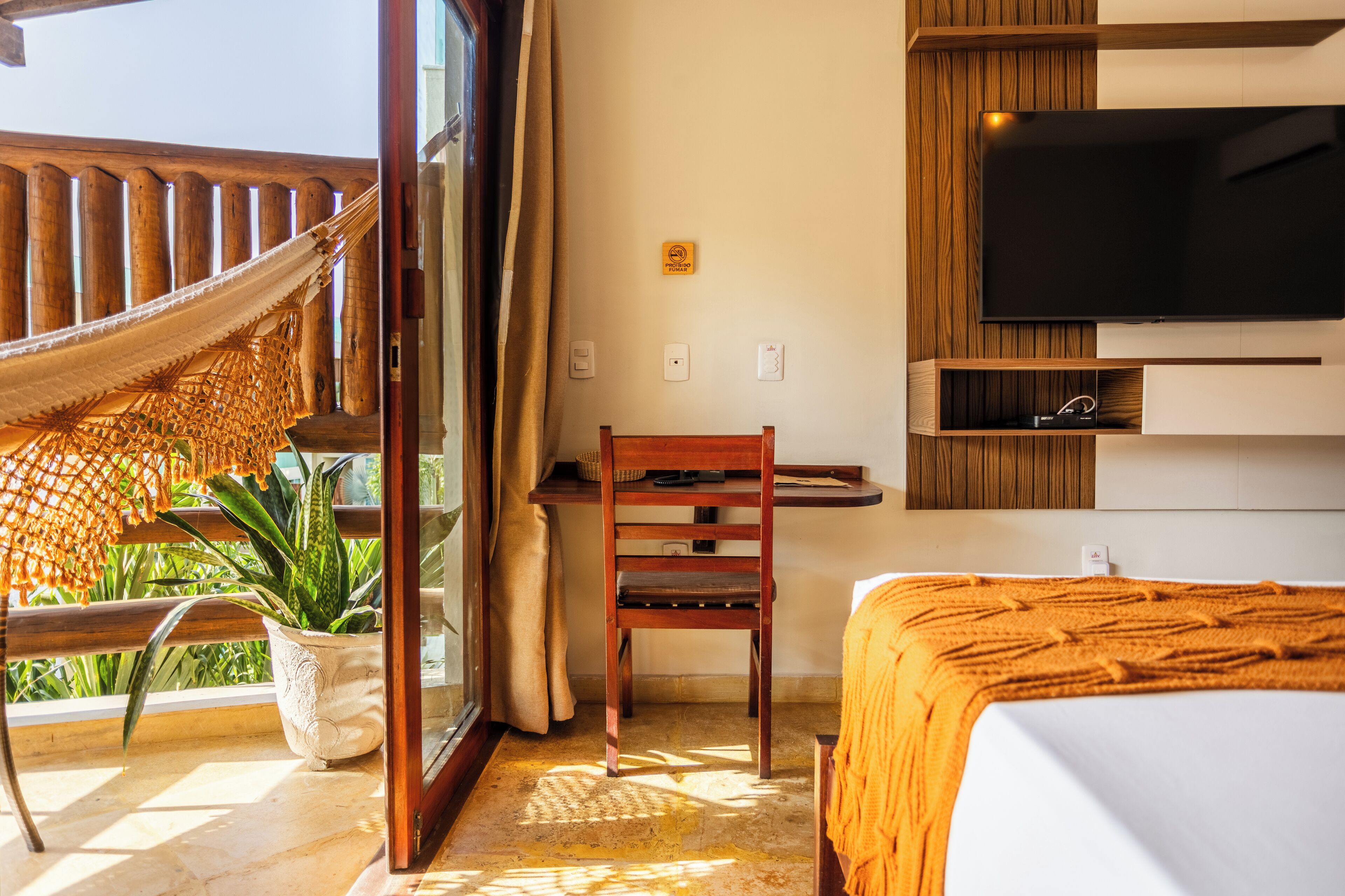 Ein Schlafzimmer mit einem Bett, einem an der Wand montierten Fernseher, einem Holzstuhl, einem kleinen Tisch und einer Hängematte vor dem Fenster.