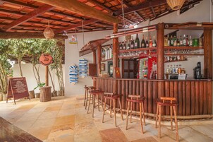 Zona de bar con taburetes de madera, una barra de madera con estantes llenos de botellas y vasos, y un cartel con texto.