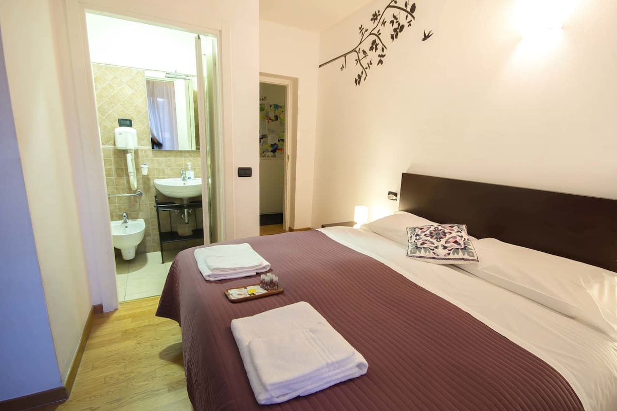Une chambre d’hôtel avec un lit, une table de chevet sur laquelle se trouve un plateau avec des articles de toilette, une salle de bain visible à travers une porte ouverte et un élément décoratif sur le mur.