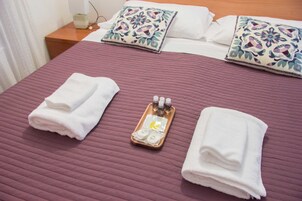 Una cama bien hecha con una colcha morada, dos toallas blancas dobladas y una bandeja con artículos de aseo.