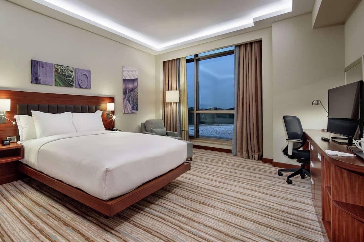 A hotel room with a large bed, a desk with a computer, a chair, a TV, and a window with curtains.