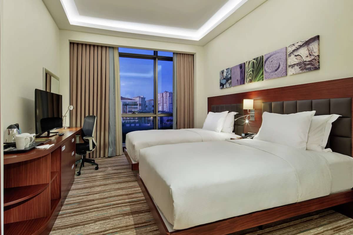 A hotel room with two beds, a desk with a TV, a chair, and a large window with a city view.