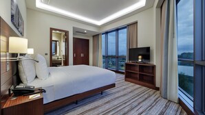 Habitación de hotel con una cama grande, un escritorio con teléfono, televisión y vistas a la ciudad a través de amplios ventanales.