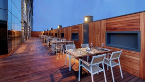 Un área de comedor al aire libre con piso de madera y sillas blancas, con mesas dispuestas para comer.